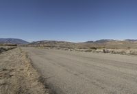 a road leading into an empty area, with grass, dirt and bushes, in the distance are mountains