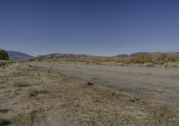 a road leading into an empty area, with grass, dirt and bushes, in the distance are mountains