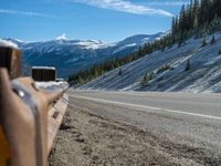 an empty highway that is snow covered in some powder and pine trees on the hill side