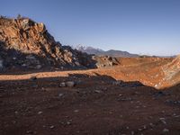 a red dirt road leading to a desert mountain with rocks and mountains in the background