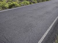 an asphalt road with a mountain in the distance with shrubs and a dirt area behind it