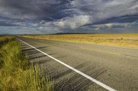 a single empty road on the prairie with dark clouds above it with a yellow stripe in between two rows