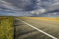 a single empty road on the prairie with dark clouds above it with a yellow stripe in between two rows