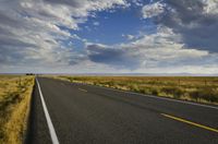 a road running along the side of a prairie with clouds and a blue sky above the road
