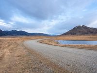 a empty road leading into a lake with mountains in the background and dry grass all around