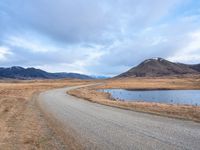 a empty road leading into a lake with mountains in the background and dry grass all around