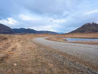 a empty road leading into a lake with mountains in the background and dry grass all around