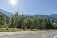 a mountain range and the sun shine brightly above the road on a sunny day with trees to the left