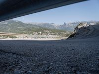 gravel road with mountains, cars, and bridge seen from behind the road to sea