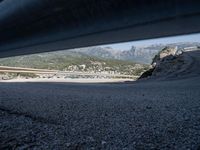 gravel road with mountains, cars, and bridge seen from behind the road to sea