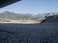 gravel road with mountains, cars, and bridge seen from behind the road to sea