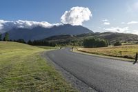 the paved roadway is surrounded by hills and grassy fields with mountains behind it and a cloudy blue sky