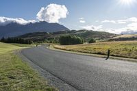 the paved roadway is surrounded by hills and grassy fields with mountains behind it and a cloudy blue sky