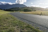 the paved roadway is surrounded by hills and grassy fields with mountains behind it and a cloudy blue sky