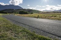 the paved roadway is surrounded by hills and grassy fields with mountains behind it and a cloudy blue sky