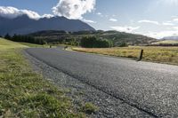 the paved roadway is surrounded by hills and grassy fields with mountains behind it and a cloudy blue sky