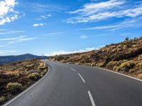 a winding highway curves with mountains in the distance and clouds above them that appear to be blue sky