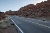 a road surrounded by dry grass with large rocky hills behind it, in the background are red rocks