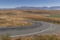 a road leading through dry grass hills with mountains in the background of the image,