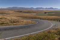 a road leading through dry grass hills with mountains in the background of the image,