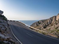 the road is winding past a cliff side area on a sunny day, with one car driving in the street