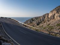 the road is winding past a cliff side area on a sunny day, with one car driving in the street