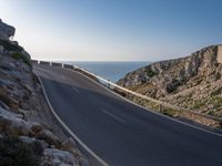 the road is winding past a cliff side area on a sunny day, with one car driving in the street