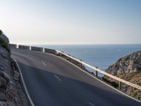 the road is winding past a cliff side area on a sunny day, with one car driving in the street