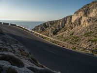 the road is winding past a cliff side area on a sunny day, with one car driving in the street