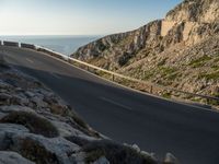 the road is winding past a cliff side area on a sunny day, with one car driving in the street