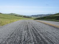 road with gravel, grass and hill in background, blue sky, and distant hills