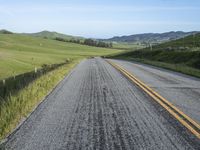 road with gravel, grass and hill in background, blue sky, and distant hills