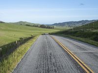 road with gravel, grass and hill in background, blue sky, and distant hills