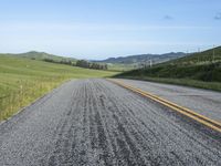 road with gravel, grass and hill in background, blue sky, and distant hills