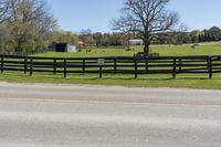 a road that has a fence on the side of it in front of trees, grass and a field in the background