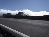 an asphalt road with a low laying line between it and the mountains with clouds in the distance