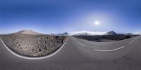 the side of a curved road with a sun behind it and mountains in the distance