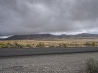 a lone asphalted road with mountains and clouds in the background behind it, along side the desert