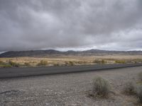 a lone asphalted road with mountains and clouds in the background behind it, along side the desert