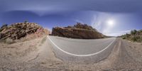 a fisheye image of a curved road by some rock formations near the mountains under a bright sky