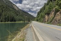 a large body of water next to a road with trees and mountains on it's sides