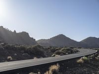 a road stretches along the side of mountains, towards a rock covered hillside with bushes