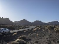 a road stretches along the side of mountains, towards a rock covered hillside with bushes