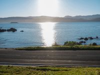 a scenic roadway leading to water next to the beach and hills in background during the sun