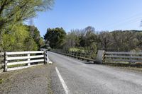 a white fence sitting next to an open road and tree filled fields under a blue sky