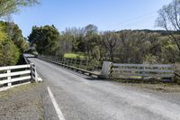 a white fence sitting next to an open road and tree filled fields under a blue sky