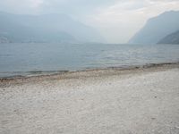 an empty bench on the beach of a lake in the background is mountains and a city in the distance