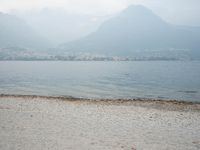 an empty bench on the beach of a lake in the background is mountains and a city in the distance