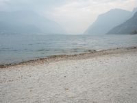 an empty bench on the beach of a lake in the background is mountains and a city in the distance