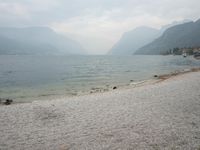 an empty bench on the beach of a lake in the background is mountains and a city in the distance